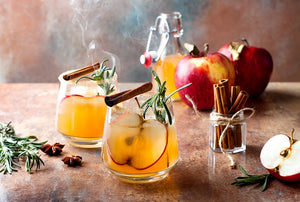 Smoked Apple Cinnamon Punch Cocktail in two glasses on a bench surrounded by garnish