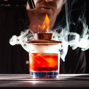 Man lighting a hardwood cocktail smoker that is sitting on a glass filled with whiskey