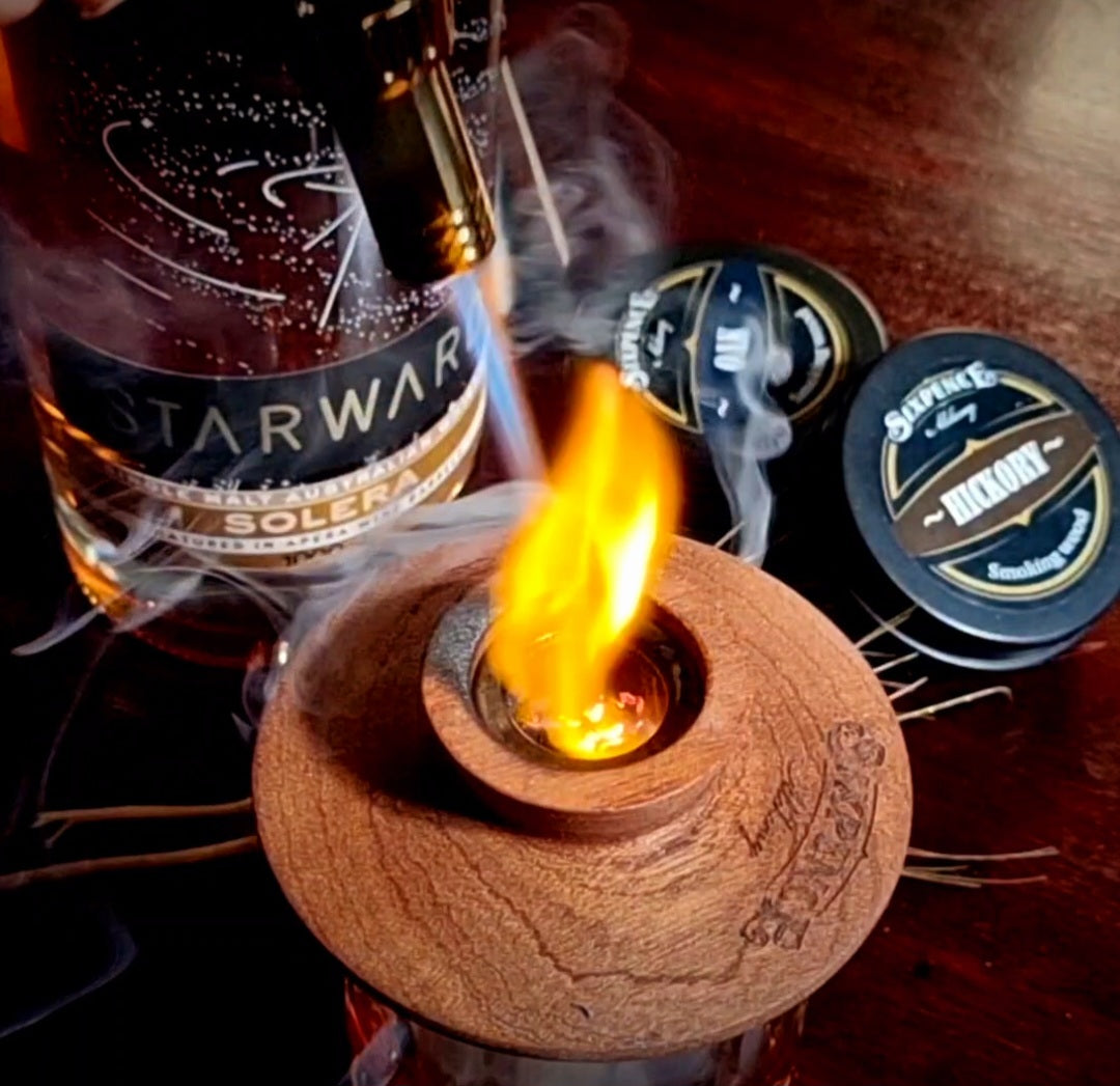 Sixpence Alchemy hardwood cocktail smoker atop a whiskey glass and alight with a flame