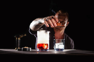 Mixologist with the Sixpence Alchemy Deluxe hardwood Cocktail Smoker, with a Mixing Glass containing a Smoked Negroni and natural woodchips and butane torch